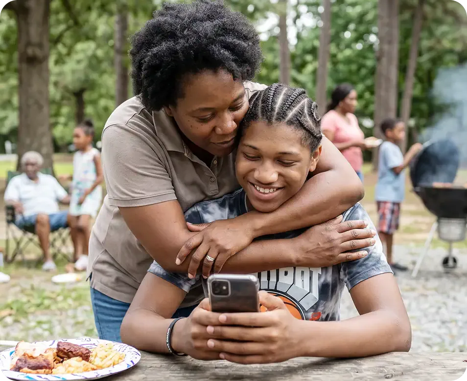 Parent hugging a smiling teenager who holds a free government iPhone outdoors